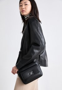 Schwarze Leder-Crossbody-Tasche mit einer strukturierten Form, silbernem Logo und strukturierten Oberfläche, gehalten von einer Person in einer schwarzen Jacke.