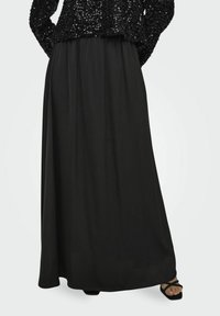 Jupe maxi noire en tissu lisse et fluide. Taille élastique en haut. Portée avec des sandales noires à brides. Top orné de sequins visible.