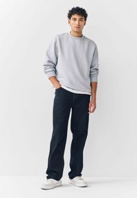 Ung mand med krøllet mørkt hår iført en grå sweatshirt, mørke jeans og hvide sneakers, stående med den ene hånd i lommen mod en hvid baggrund.