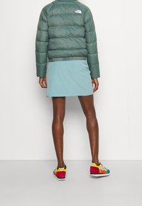 Giacca verde gonfia, gonna blu chiaro e sneakers multicolori. La giacca ha un logo sulla schiena e presenta un cinturino a coste.