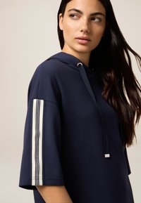 Sudadera con capucha navy de manga corta; presenta dos franjas verticales blancas en la manga izquierda, fabricada con un tejido suave.