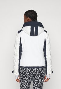Chaqueta acolchada blanca y marino con un alto cuello, silueta ajustada y textura acolchada. Presenta detalles reflectantes y mangas en contraste.