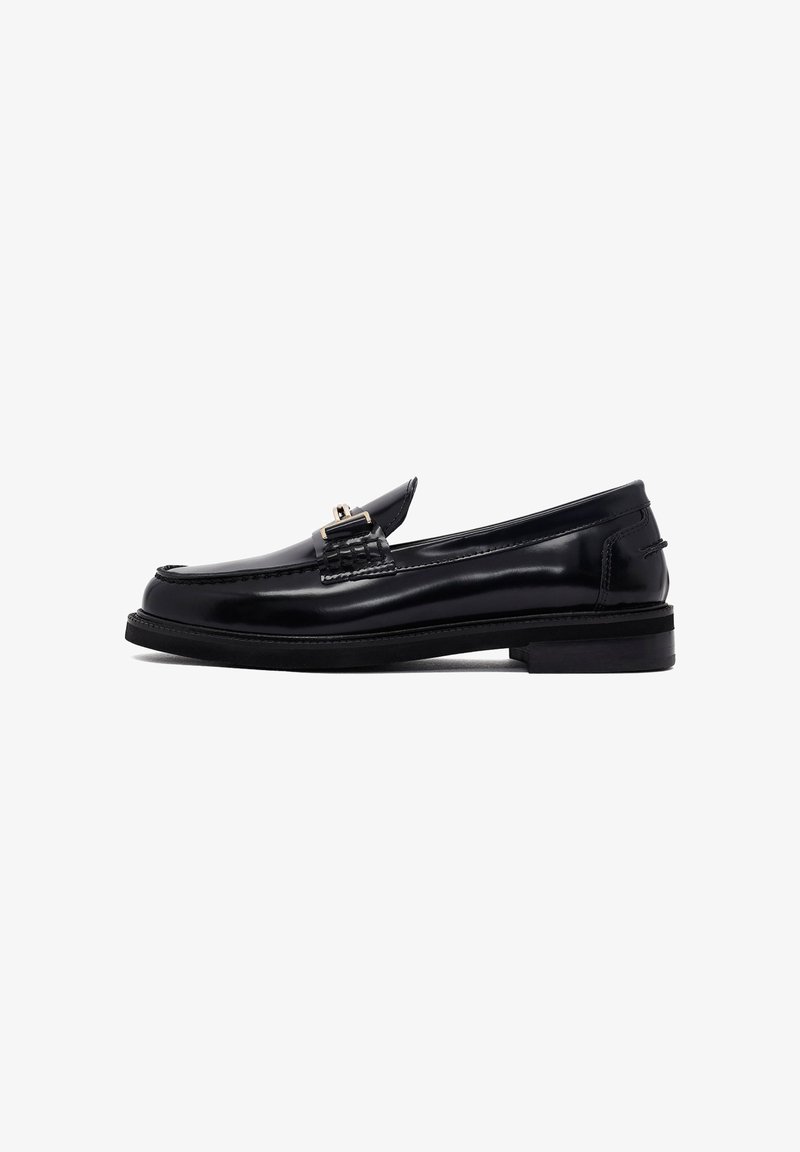 Mocassin en cuir noir avec un talon plat, présentant un détail tissé et une quincaillerie décorative à l'avant, texture lisse et silhouette classique.