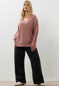 Vaaleanpunainen neulepusero, jossa on V-pääntie, pitkät hihat ja napit hihansuissa, yhdistettynä leveälahkeisiin mustiin housuihin ja valkoisiin tenniqueen.