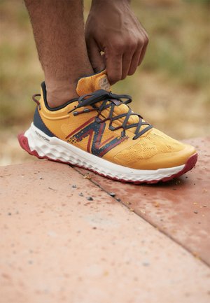 Person bindet graue Schnürsenkel an einem gelb-orangefarbenen Laufschuh mit weißer Sohle und der Aufschrift Fresh Foam auf einer Außenfläche.