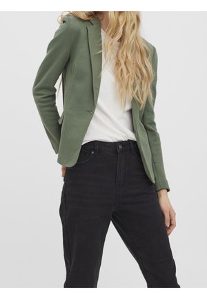 Frau trägt ein tailliertes olivgrünes Blazer, weißes Oberteil und hoch taillierte schwarze Jeans vor einem einfarbigen hellgrauen Hintergrund.