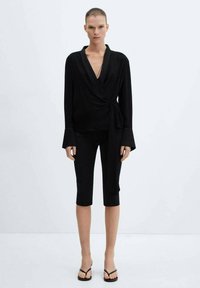 Top nero avvolgente con scollatura a V profonda, maniche a campana lunghe e vita legata, abbinato a pantaloni neri aderenti e cropped e sandali neri.