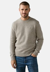 Lys grå striktrøje med rund hals og ribbede mansjetter, prydet med et lille broderet logo på brystet, som er parret med blå denim jeans.