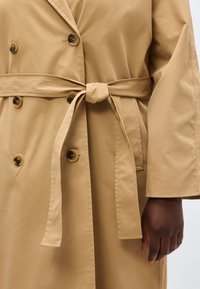 Trench-coat camel à double boutonnage avec des boutons en écaille, ceinturé à la taille, porté par une personne à la peau foncée et au bras détendu.
