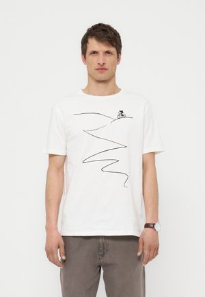 Uomo che indossa una t-shirt bianca con un disegno a linee nere di un ciclista che scende una collina tortuosa, abbinata a pantaloni grigi e un orologio.