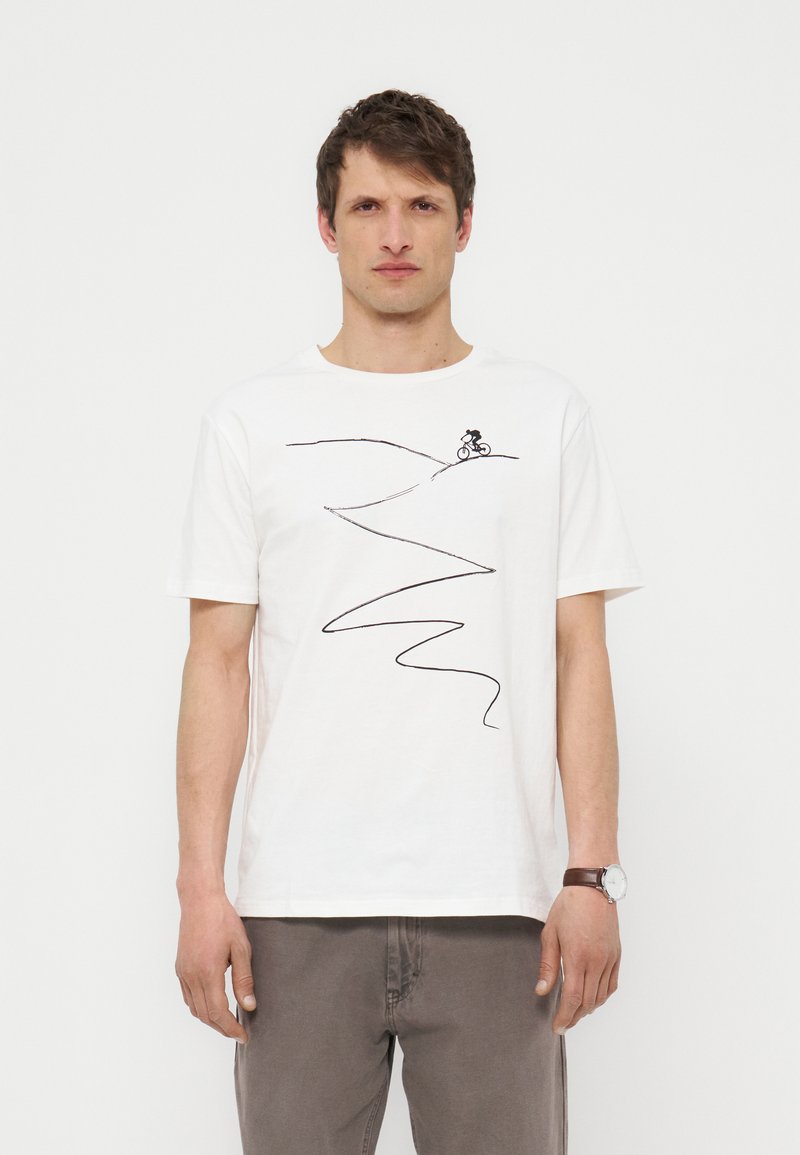 Άνδρας που φοράει λευκό μπλουζάκι με μαύρο σχέδιο ενός ποδηλάτη που κατεβαίνει έναν καμπυλωτό λόφο, συνδυασμένο με γκρι παντελόνι και ρολόι.