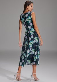 Blumenkleid in Midi-Länge mit navyblauem Grundton, das mit weißen und grünen Blumen verziert ist. Ärmelloses Design, weicher Stoff und eine fließende Silhouette. Silberne Absätze.