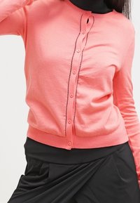 Korallrosa kofta tillverkad av mjukt stickat tyg, med rund halsringning, knappstängning och dekorativa tråddekorationer längs knappslån.