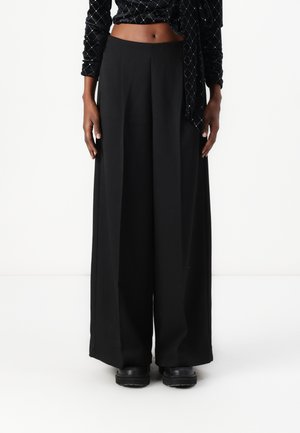 Pantalons larges noirs avec une texture lisse, taille haute et une coupe ample. Stylés avec un haut ajusté et des chaussures noires.
