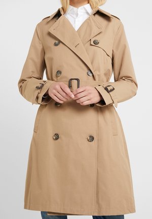 Beige double-breasted trench coat with belt and buttoned straps on sleeves worn over white shirt on a person.