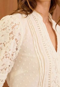 Blusa bianca con pois testurizzati e dettagli in pizzo sulle maniche a sbuffo. Presenta pieghe verticali e un design a V. Tessuto leggero.