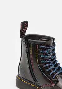 Botín de cuero negro con brillo, con costuras multicolores, cordones arcoíris, cremallera lateral y una lengüeta en el talón.