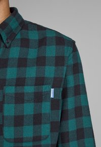 Camisa a cuadros verde y negra con cuello abotonado, un bolsillo en el pecho y una pequeña etiqueta azul en la costura del bolsillo. Tejido texturizado.