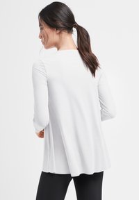 Hvid langærmet bluse med en løs pasform, rund halsudskæring og plisseret rygdesign, parret med sorte leggings. Glat stoftekstur.