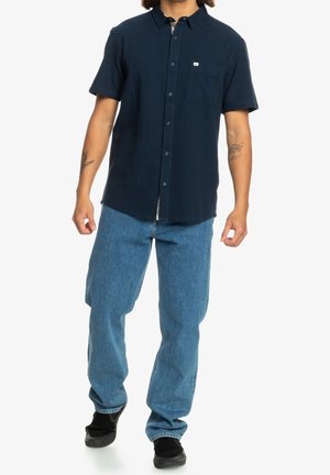 Homme portant une chemise bleu marine à manches courtes avec des boutons, un jean bleu clair et des chaussures noires, debout devant un fond blanc.