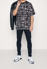 Černé tričko s kontrastním bílým textem a logy, spárované s černými kalhotami a stříbrnými teniskami. Oblečení doplňuje strukturovaná kožená bunda.