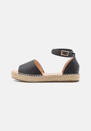Sandale espadrille en cuir noir à bout ouvert avec bride cheville et boucle sur semelle plateforme en jute tissé sur fond blanc.
