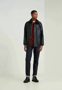 Homem em pé a usar um casaco encerado escuro, suéter bordô, jeans escuros com as pernas arregaçadas e botas pretas de atar, contra um fundo simples.