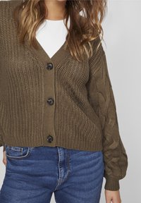 Kvinna som bär en brun stickad knappar kofta över en vit skjorta, tillsammans med blå denimjeans, mot en neutral bakgrund.