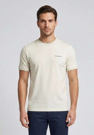 Uomo con capelli castani corti che indossa una maglietta beige chiaro semplice e pantaloni blu scuro, in piedi davanti a uno sfondo grigio chiaro uniforme.