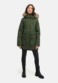 Parka verde con cappuccio foderato in pelliccia, chiusure a zip e bottoni, due tasche frontali e vita regolabile con coulisse, indossata sopra jeans neri e stivali.