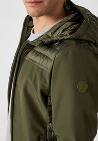 Giacca isolante verde oliva con cappuccio, caratterizzata da una combinazione di texture lisce e trapuntate. Panno con logo sul braccio sinistro, dettaglio con zip visibile.
