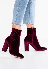 Burgundské sametové kotníkové boty s elegantním designem, špičatou špičkou a 7,5 cm blokovým podpatkem; obsahují jemné nařasení a boční zip.