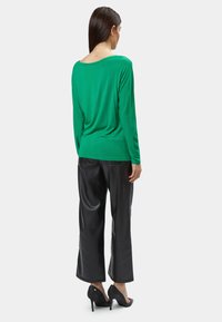 Grünes langärmliges Oberteil, kombiniert mit schwarzen, weit geschnittenen Hosen und schwarzen High Heels; weicher Stoff mit lockerer Passform und einem runden Ausschnitt hinten.