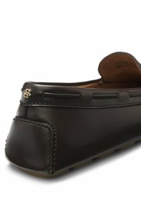 Gros plan sur un mocassin en cuir noir avec des coutures détaillées, une semelle en caoutchouc et un petit logo doré sur le talon.