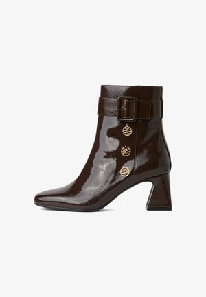 Glanzende donkerbruine enkelboot met blokhak, spitse neus, brede riem met gesp en drie decoratieve gouden knoppen aan de zijkant.