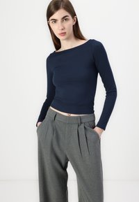 Top ajustado de manga longa azul-marinho com decote barco, combinado com calças cinzas plissadas de cintura alta. O tecido parece suave e elástico.