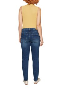 Schlanke dunkelblaue Jeans mit glatter Textur, Fünf-Taschen-Design und kontrastierenden Nähten. Getragen mit einem ärmellosen gelben Oberteil.