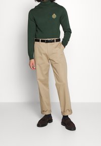 Grüner Rollkragenpullover mit einem gestickten Emblem, kombiniert mit beigen Chinos und braunen Wildlederschuhen, die eine lockere Passform und ein einfaches Design zeigen.