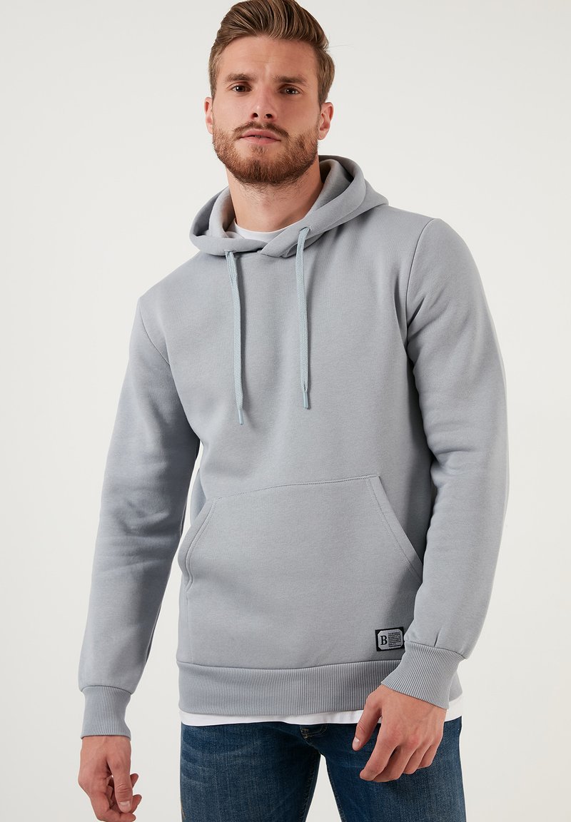 Sudadera gris claro de tejido suave con un bolsillo frontal, capucha ajustable con cordón, puños de canalé y un parche de logo en el dobladillo inferior.