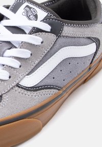 Γκρι και μαύρο αθλητικό παπούτσι Vans με λευκή λωρίδα στο πλάι, λευκά κορδόνια και σόλα από καουτσούκ χρώματος καραμέλας, που δείχνει λε πτομερείς ραφές και το λογότυπο στη γλώσσα του παπουτσιού.