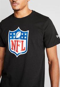 Hombre que lleva una camiseta negra con un gran logo de la NFL en azul, rojo y blanco, y el logo blanco de New Era en la manga derecha.