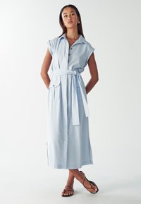 Donna in piedi con le braccia dietro la schiena, indossa un abito azzurro chiaro senza maniche e con bottoni, cintura annodata, tasche frontali e sandali neri con cinturini.