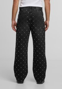 Melnas platās kājas džinsus ar izšūtu dimanta rakstu un nelieliem kniedēm, valkā ar baltu topu, redzami no aizmugures.