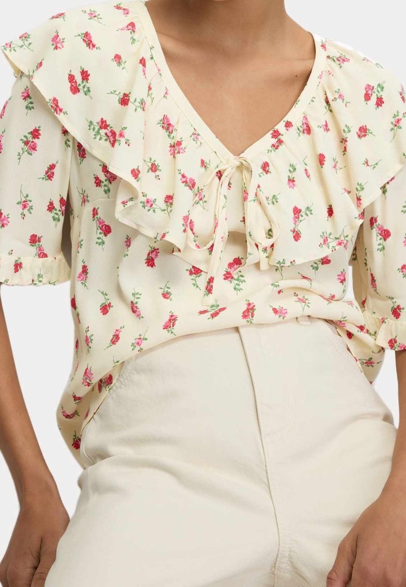 Femme portant un chemisier crème à imprimé floral rouge, col volanté, manches courtes bouffantes et lien à l'avant, associé à un pantalon crème taille haute.