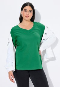 Zielona koszulka z krótkim rękawem i dekoltem w serek, wykonana z gładkiej tkaniny, zestawiona z białą kurtką. Modelka pozuje z rękami na biodrach na białym tle.