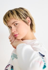 Hvit sweatshirt med fargerike grafiske design, myk tekstur, rund hals og avslappet passform. Kort, lagdelt blond frisyre.