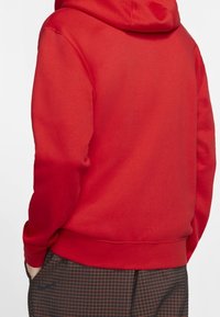 Person mit einem schlichten roten Kapuzenpullover, die Kapuze aufgesetzt, und schwarzen Hosen mit einem roten Karomuster, die Hände in den Taschen, von hinten gezeigt.