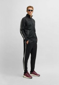 Black athletic tracksuit with white side stripes, zippered jacket, slim-fit pants, and maroon sneakers. Fabric appears smooth and textured.