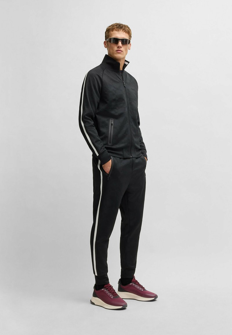 Black athletic tracksuit with white side stripes, zippered jacket, slim-fit pants, and maroon sneakers. Fabric appears smooth and textured.