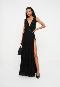 Vestido preto com um topo em V profundo decorado com contas e uma saia plissada fluida com uma fenda alta. Acessórios incluem uma pequena carteira preta.